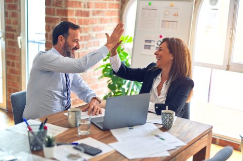 Ein Mann und eine Frau in Geschäftskleidung geben sich im Büro ein High-Five. Sie sitzen an einem Schreibtisch mit einem Laptop, Papierdokumenten und persönlichen Gegenständen, lächeln und zeigen Zeichen von Erfolg und positiver Zusammenarbeit.